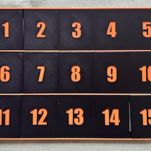 May include: A rectangular black grid organizer featuring three horizontal rows, each containing five individual compartments. Each compartment displays a raised orange number, sequentially ordered from one to fifteen across the entire grid.