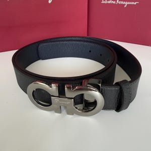 Puede incluir: Un cinturón de cuero negro texturizado con una hebilla de metal plateado que presenta el logotipo Gancini y el nombre de la marca Salvatore Ferragamo grabado en el centro. El cinturón incluye una trabilla de cuero y orificios de ajuste.