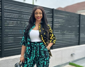 May include: A woman's ensemble featuring a patterned jacket with black, yellow, and teal accents, paired with matching pants. The outfit is complemented by a white top and a black handbag. The background includes a black fence and green grass.