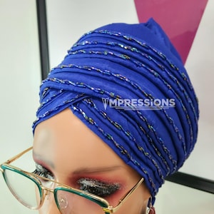 May include: A blue turban with iridescent beads wrapped around it. The turban is worn by a person with red eyeshadow and gold-rimmed glasses with green lenses. The person is wearing red tassel earrings.