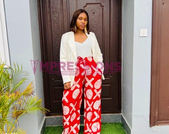 Puede incluir: Una mujer con una blazer blanca, un top blanco y pantalones estampados rojos y blancos. Los pantalones tienen un diseño de pierna ancha y un lazo en la cintura. El fondo incluye una puerta marrón y una alfombra verde.