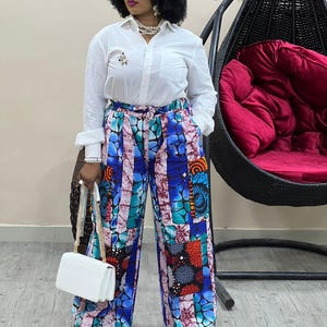 May include: A white button-down shirt paired with wide-leg patterned pants in blue, teal, and orange. A white purse with a gold chain strap and black and white sneakers complete the outfit. The pants feature a unique patchwork design.