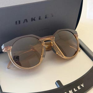 May include: Oakley sunglasses featuring round, brown-tinted lenses with a two-tone frame design. The upper portion of the frame is dark brown, while the lower portion is a translucent amber color. The glasses are displayed in front of an Oakley box.