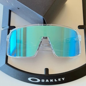 May include: Clear frame sunglasses featuring a single-lens shield design with a reflective green and blue gradient finish. The eyewear displays the Oakley brand name on the lens and is positioned in front of a black branded display case.