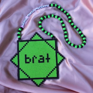 May include: A neon green and black beaded necklace with a pendant that reads "brat". The pendant is a pixelated design with a starburst shape. The necklace has a black and green striped pattern.