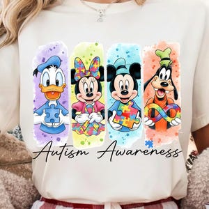 Pode incluir: Camiseta branca com os personagens de desenhos animados Pato Donald, Minnie, Mickey e Pateta, cada um com uma peça de quebra-cabeça ou um símbolo de infinito. O texto "Autism Awareness" está escrito em itálico preto.