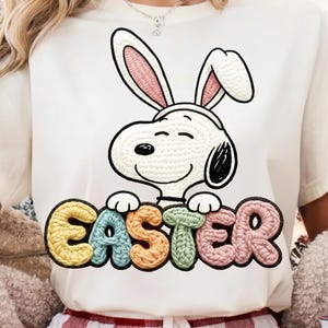 以下が含まれることがあります： クリーム色のTシャツ。ウサギの耳をつけたスヌーピーの漫画のキャラクターと、カラフルで質感のある文字で「EASTER」という文字が描かれています。デザインはTシャツの中央に配置され、お祝いのイースターをテーマにしています。