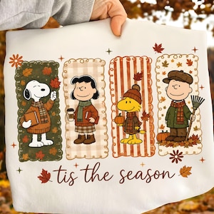May include: A white sweatshirt featuring a graphic of Snoopy, Lucy, Woodstock, and Charlie Brown dressed in autumn clothing. The characters are in four vertical panels with fall leaves and the text 'tis the season' printed in brown script below.