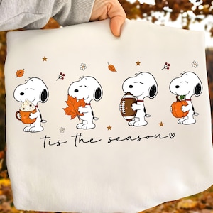 May include: A white sweatshirt featuring a graphic of Snoopy in four poses: holding a pumpkin mug, an orange leaf, a football, and a pumpkin. The text "tis the season" is printed below the characters, accompanied by small stars and floral icons.