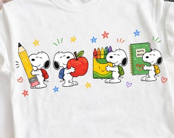 May include: A white short-sleeved t-shirt featuring a graphic of the cartoon character Snoopy in four poses, holding a large yellow pencil, a red apple, a box of colorful crayons, and a green notebook labeled NOTE BOOK, surrounded by small stars and hearts.