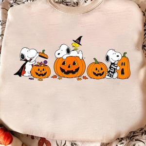 May include: A beige crewneck sweatshirt featuring a graphic print of Snoopy and Woodstock characters celebrating Halloween. The design includes multiple orange jack-o'-lanterns, candy, and the characters wearing costumes like a vampire cape and witch hat.