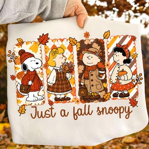 Könnte beinhalten: Ein weißes Sweatshirt mit einer Grafik von Snoopy und drei Peanuts-Figuren in herbstlicher Kleidung, umgeben von Herbstblättern. Die Figuren sind in vier vertikalen Feldern angeordnet, darunter steht der Text Just a fall snoopy.