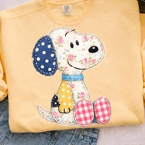 May include: A light yellow sweatshirt featuring a Snoopy applique. The Snoopy design includes a blue polka dot ear, floral print body, and pink gingham feet. The sweatshirt has a crew neck and long sleeves.