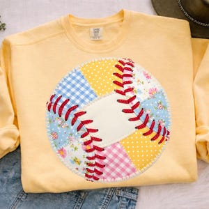 Peut inclure: Un sweat-shirt jaune clair avec un motif de baseball. Le baseball est fait de tissu patchwork dans divers motifs, dont du vichy bleu, des motifs floraux et des pois jaunes. Des coutures rouges détaillent les coutures du baseball.