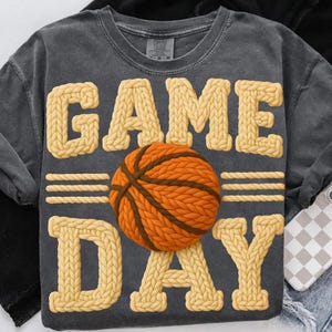 May include: Dark gray t-shirt with the words "GAME DAY" in a knitted style, featuring a basketball in the center. The basketball is orange and brown. The shirt has a vintage, washed look.