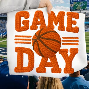 May include: A white sign with the words "GAME DAY" in orange knit letters. A basketball with brown knit details is in the center. The sign is held up at a sporting event.
