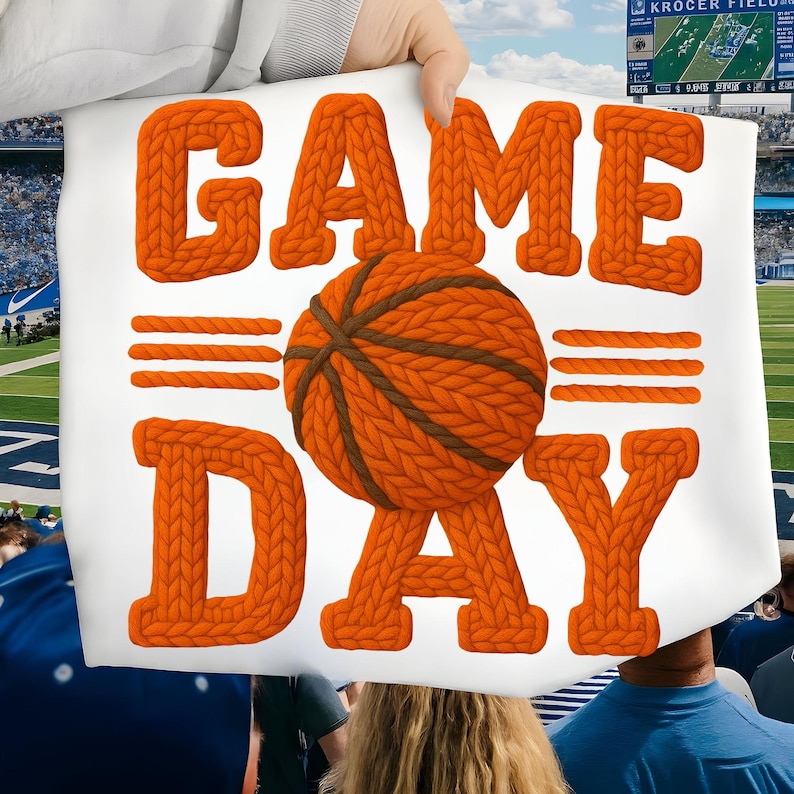 May include: A white sign with the words "GAME DAY" in orange knit letters. A basketball with brown knit details is in the center. The sign is held up at a sporting event.