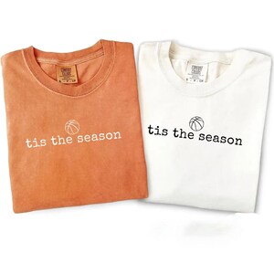 Puede incluir: Se muestran dos camisetas, una naranja y otra blanca. Cada camiseta tiene la frase "tis the season" impresa en blanco o negro, con un gr&aacute;fico de baloncesto encima del texto. Las camisetas est&aacute;n dobladas.