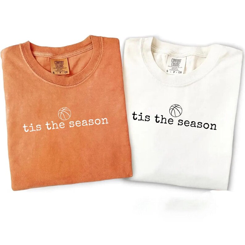 Puede incluir: Se muestran dos camisetas, una naranja y otra blanca. Cada camiseta tiene la frase "tis the season" impresa en blanco o negro, con un gr&aacute;fico de baloncesto encima del texto. Las camisetas est&aacute;n dobladas.