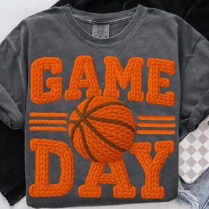 May include: A gray, long-sleeved t-shirt with the words "GAME DAY" in orange, textured, knitted letters. A basketball with orange and brown knitted details is centered on the shirt. The shirt is a Comfort Colors brand.
