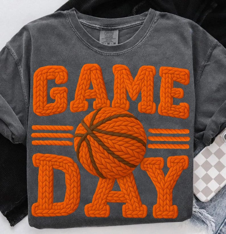 May include: A gray, long-sleeved t-shirt with the words "GAME DAY" in orange, textured, knitted letters. A basketball with orange and brown knitted details is centered on the shirt. The shirt is a Comfort Colors brand.