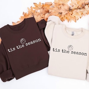 Puede incluir: Dos sudaderas, una marr&oacute;n y otra crema, con el texto "tis the season" y un gr&aacute;fico de baloncesto. El texto est&aacute; en una fuente sencilla. Las sudaderas est&aacute;n dobladas y expuestas sobre una superficie con hojas de oto&ntilde;o.
