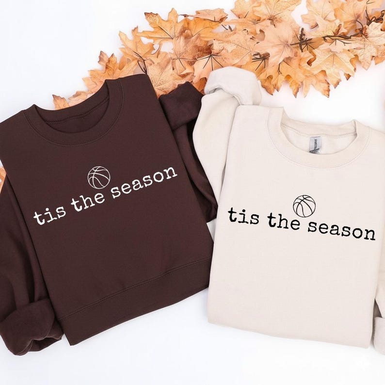 Puede incluir: Dos sudaderas, una marr&oacute;n y otra crema, con el texto "tis the season" y un gr&aacute;fico de baloncesto. El texto est&aacute; en una fuente sencilla. Las sudaderas est&aacute;n dobladas y expuestas sobre una superficie con hojas de oto&ntilde;o.