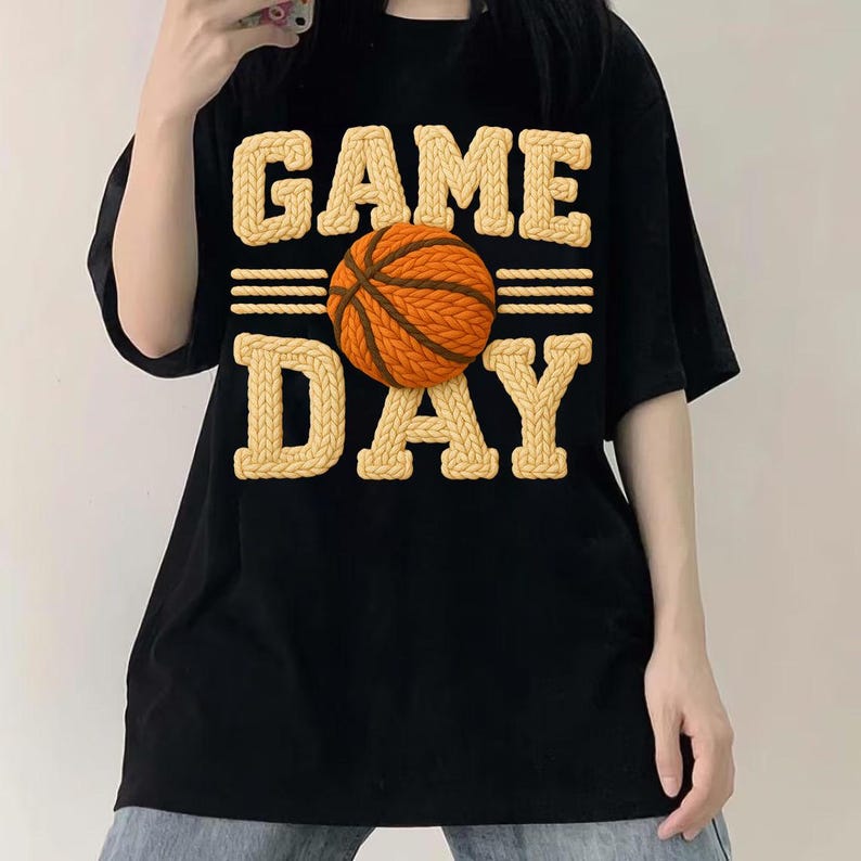 May include: Black t-shirt with the words "GAME DAY" in a textured, light tan font, with a basketball in the center. The basketball is orange and brown. The shirt is a casual style.