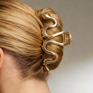 Puede incluir: Una pinza de pelo de metal dorado con forma de serpiente enroscada. El accesorio presenta una superficie pulida y reflectante con un diseño serpentino detallado y sinuoso que sujeta el cabello en un moño. La pinza tiene un mecanismo de resorte.
