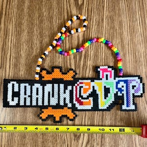 May include: A black pixel art sign with the word "CRANKCUP" in white and colorful letters. The sign is adorned with orange pixel art accents and a beaded necklace in various colors. A measuring tape is at the bottom.