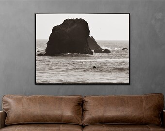 May include: A black and white photograph of a large rock formation in the ocean. The rock is in the foreground, with waves crashing around it. A lone figure is seen surfing in the distance.
