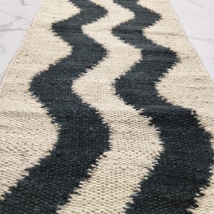 May include: A rectangular rug featuring a cream-coloured base with two wavy, dark grey stripes running along its length. The rug has a textured, woven appearance, suitable for a hallway or entryway.