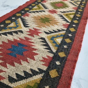 May include: A rectangular jute runner rug with a geometric pattern. The rug features a central design with red, blue, green, and yellow shapes on a beige background. It has a black border with yellow accents and a red edge.