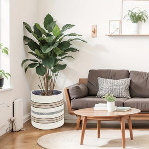 Puede incluir: Una planta en maceta grande con hojas verdes anchas en una maceta tejida blanca con rayas horizontales en beige, azul marino y gris. La maceta está en un salón con un sofá marrón y una mesa de centro de madera. La maceta mide unos 40 cm de diámetro.