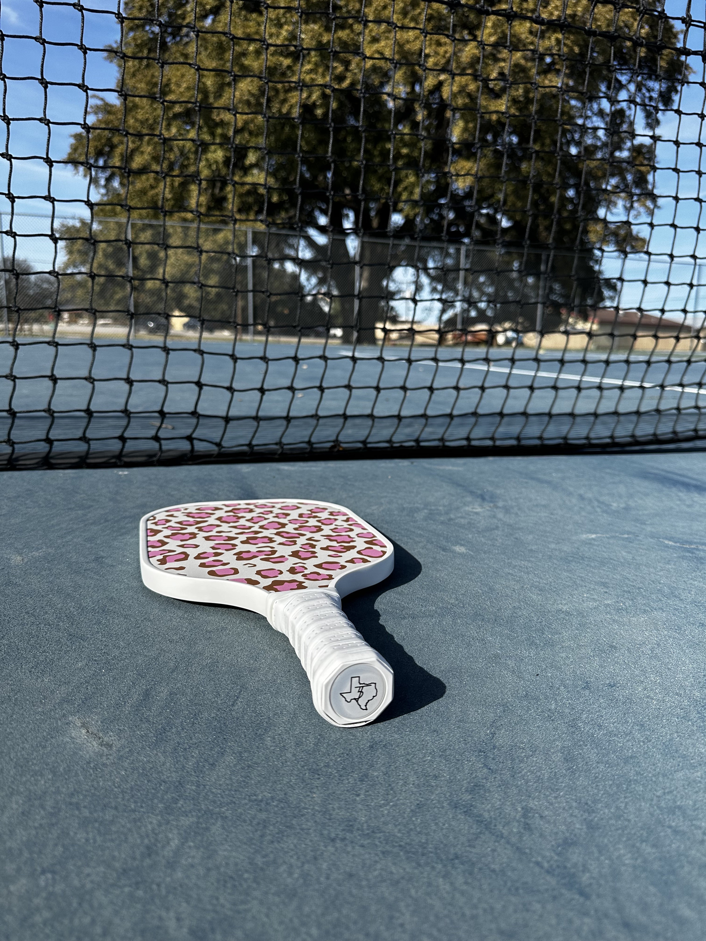 May include: White pickleball paddle with a pink and brown leopard print design. The paddle has a white handle with a Texas-shaped logo.