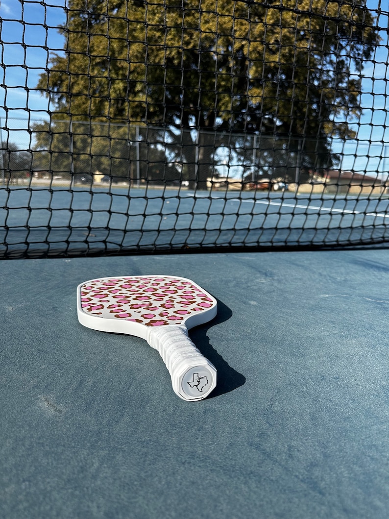 May include: White pickleball paddle with a pink and brown leopard print design. The paddle has a white handle with a Texas-shaped logo.