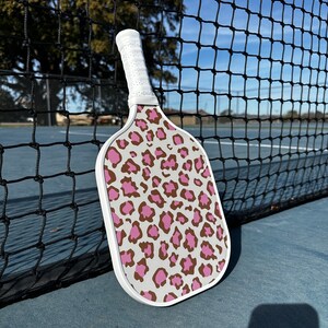 May include: A white pickleball paddle with a pink and brown leopard print design. The paddle is leaning against a black tennis net.