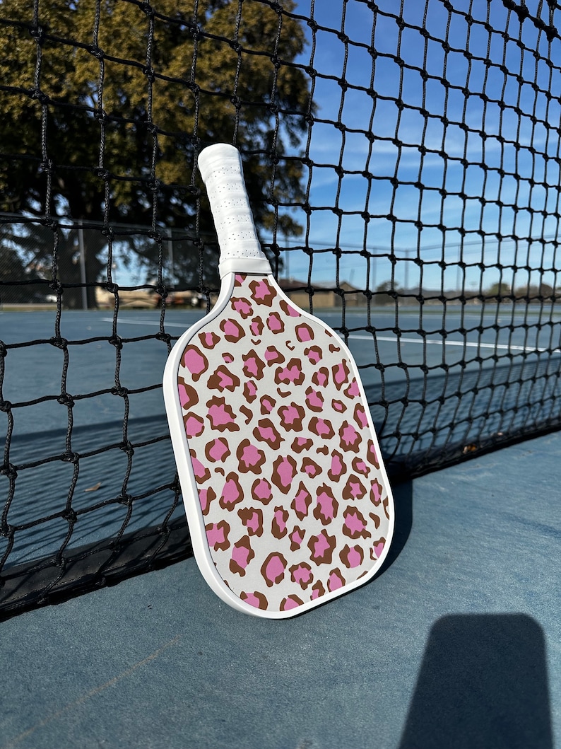 May include: A white pickleball paddle with a pink and brown leopard print design. The paddle is leaning against a black tennis net.