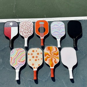 May include: Nine different patterned pickleball paddles. The paddles are all white with different colored designs. The designs include a Texas flag, a leopard print, a guitar, a swirl pattern, a solid black paddle, a rainbow swirl, a daisy pattern, an orange swirl, and a solid white paddle.