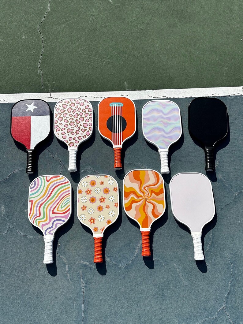 May include: Nine different patterned pickleball paddles. The paddles are all white with different colored designs. The designs include a Texas flag, a leopard print, a guitar, a swirl pattern, a solid black paddle, a rainbow swirl, a daisy pattern, an orange swirl, and a solid white paddle.