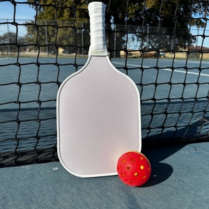 May include: A light purple pickleball paddle with a white grip and a red pickleball with yellow holes. The paddle and ball are on a blue court, with a black net in the background.