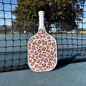 May include: A white pickleball paddle with a pink and brown leopard print design. The paddle is leaning against a black net.