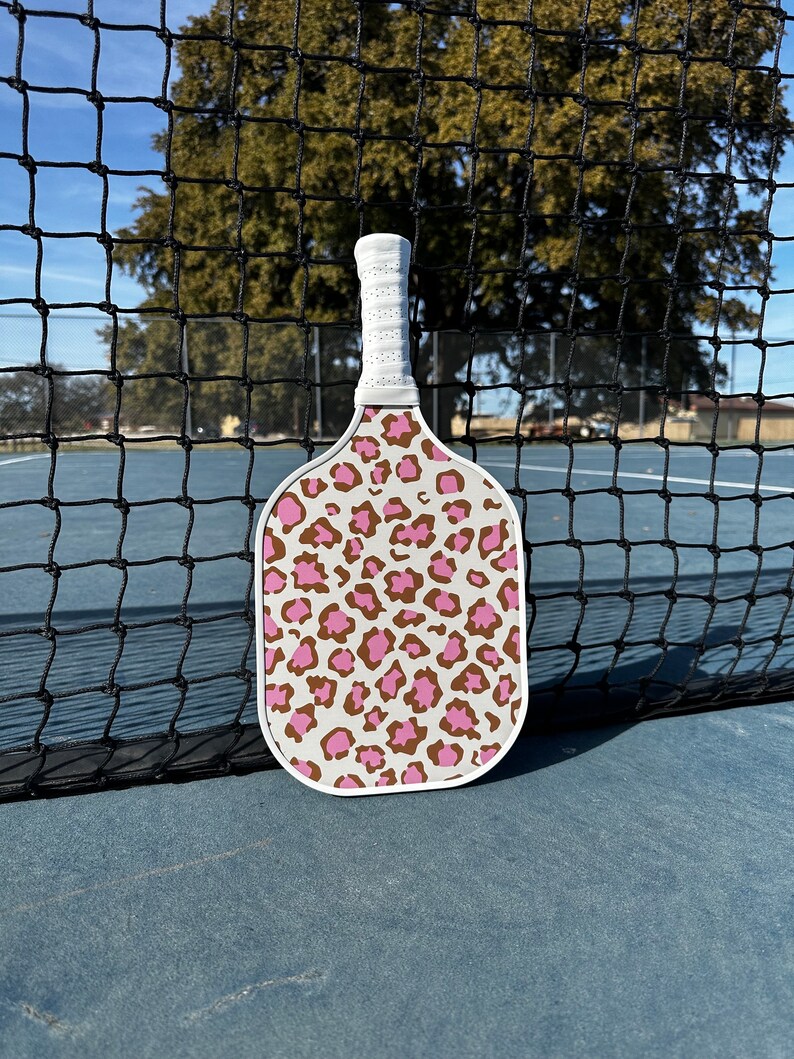 May include: A white pickleball paddle with a pink and brown leopard print design. The paddle is leaning against a black net.
