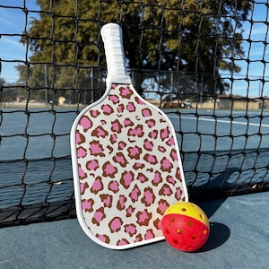 May include: A white pickleball paddle with a pink and brown leopard print design. A red and yellow pickleball is next to the paddle.