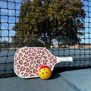May include: A white pickleball paddle with a pink and brown leopard print design. A yellow pickleball with red accents is next to the paddle.