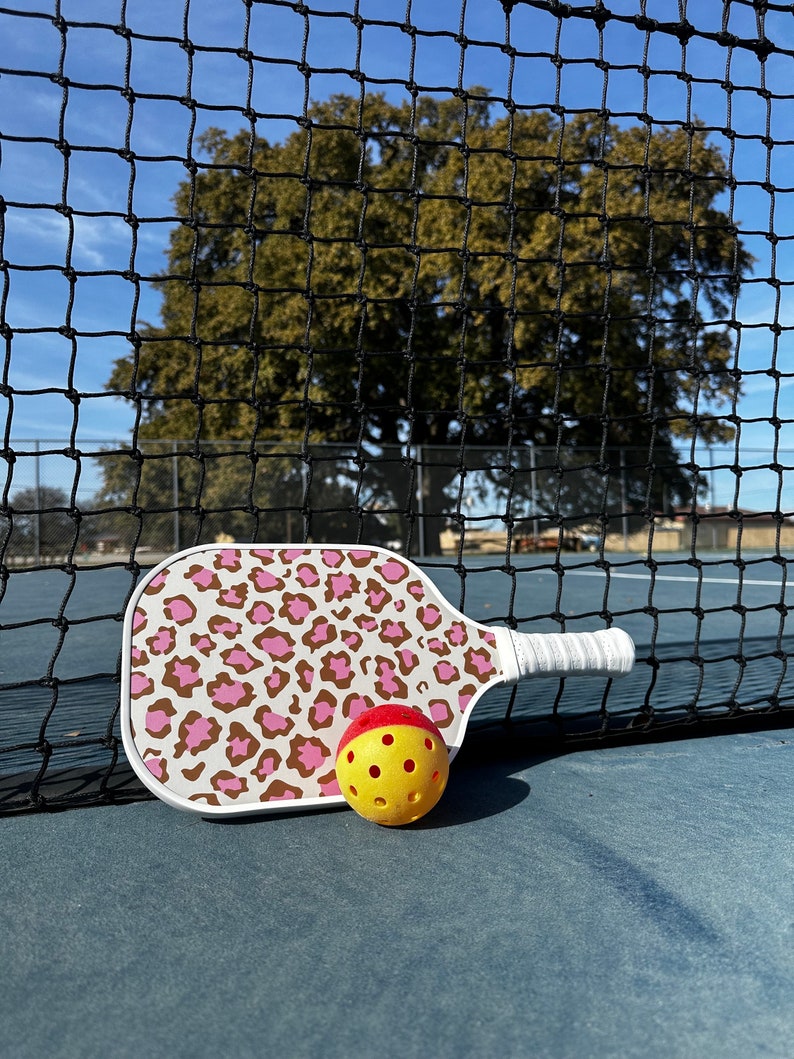 May include: A white pickleball paddle with a pink and brown leopard print design. A yellow pickleball with red accents is next to the paddle.