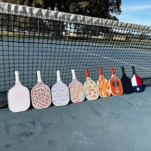 May include: Nine different patterned pickleball paddles lined up on a blue tennis court. The paddles have various designs, including animal print, swirls, and abstract patterns.