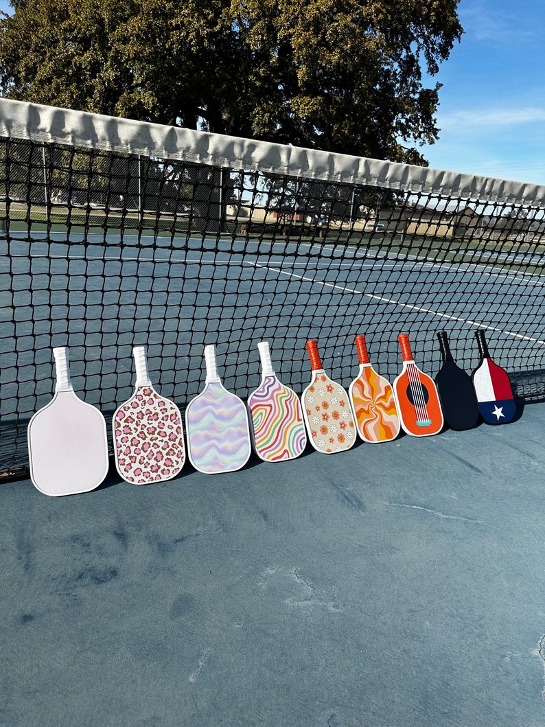 May include: Nine different patterned pickleball paddles lined up on a blue tennis court. The paddles have various designs, including animal print, swirls, and abstract patterns.