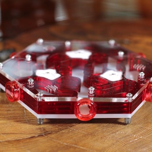 May include: A clear and red hexagonal ant farm with a white base and silver fixings. The transparent design allows for easy viewing of the ant colony. The red accents and rounded corners give it a modern look.
