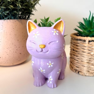 May include: A lavender cat-shaped planter featuring a hand-painted white daisy pattern across its body. The cat has gold-painted inner ears and a gold nose, with a smooth, matte finish. The top of the cat's head is open to hold a small plant.